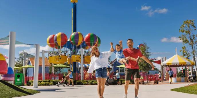 A mother and father swinging their young son by his arms while walking in front of a Peppa Pig balloon ride at a theme park