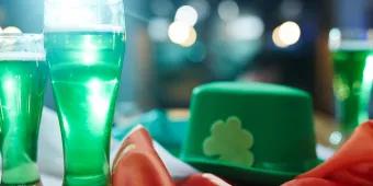 A glass full of green liquid next to a green hat with a shamrock and an Irish flag