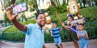 A father and three children taking a selfie posing with lightsabers in front of the Star Wars statues