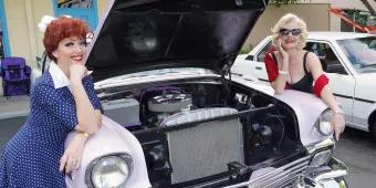 Two women wearing vintage clothes and make-up leaning on a classic car