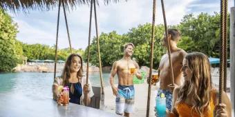 Two women sat on swing seats and two men standing behind them on a beach, all holding cocktails