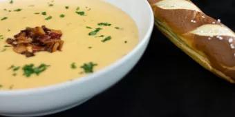 A bowl of bacon topped beer cheese soup with a pretzel roll