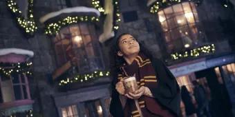 A young woman wearing a scarf and wizard robes drinking Butterbeer in front of decorated buildings