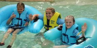 Two children and one adult wearing life jackets and waving at the camera while sitting in water