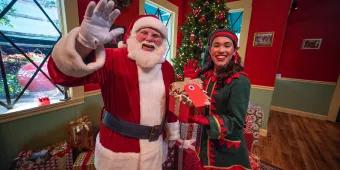 Santa waving at the camera standing next to one of his elves who is holding a present. They are standing in front of a Christmas tree