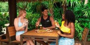 A man and two women sat around a table with two pizzas on it. The women are holding wine glasses and there is a glass of beer in front of the man