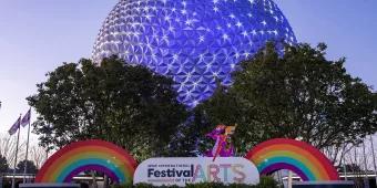 Festival of the Arts at EPCOT Spaceship Earth lit up in the early evening. In front of it is a large paint tube and a Festival of the Arts sign next to two rainbows