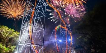 Large brightly coloured fireworks in the night sky above a tall and steep rollercoaster