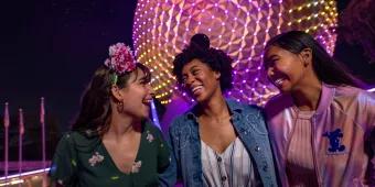 Large circular structure lit in pink and orange lighting with a group of girl friends stood in front of Spaceship Earth wearing mickey ears.