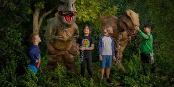 Dino Mania at Dubai Parks and Resorts 4 children standing in some foliage with two dinosaur animatronics
