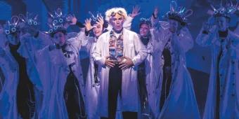Doc Brown wearing a white lab coat standing centre stage in front of dancers also wearing lab coats