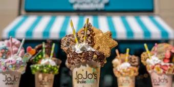 5 large milkshakes topped with cream, marshmallows, candy floss and sprinkles, in front of  sign reading JoJo's ShakeBar