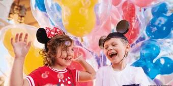 A young girl wearing a headband with Minnie Mouse ears and a young boy wearing a hat with Mickey Mouse ears standing in front of multi-coloured balloons