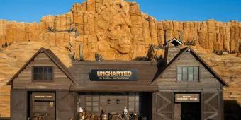 A wooden barn-like building in front of sandy cliffs with a carved Aztec face in the centre