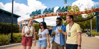 A group of four adults walking away from a roller coaster carrying pints of beer