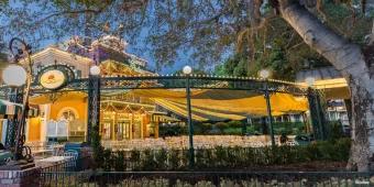 The exterior of a restaurant with white chairs and tables covered by a green and yellow striped awning and green ironwork