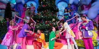 Performers dressed as Whos from the Grinch wearing bright coloured clothes and exchanging gifts in front of a large Christmas tree