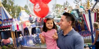 A man holding a young girl who is carrying a balloon standing in front of Dumbo the Flying Elephant