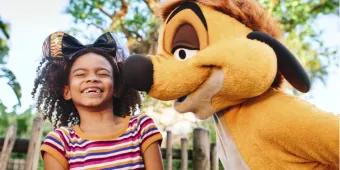 A girl smiling with eyes closed in front of the Tree of Life wearing glittery gold Mickey ears with a black bow on them. Stood next to her is Timone from the Lion King.