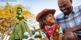 A young boy dressed as a cowboy smiling up at his dad. They are both sat in front of a topiary shaped like Bo Peep from Toy Story
