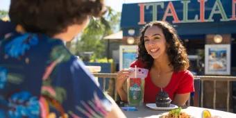 A woman eating a cupcake and drinking a cocktail from a pouch sitting across a table from someone