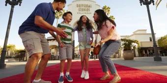 Family jumping on the red carpet in front of the large white stone entrance sign with Universal Studios Hollywood logo on. The family has dad on the right, one girl and one boy in the middle jumping and the mum crouched on the right.