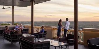 Capa in the Four Seasons hotel Terrace with brown chairs and glass tables on, there are two groups of two people sat to the left of the image and a couple stood up overlooking the view of the terrace.