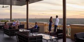 Terrace with brown chairs and glass tables on, there are two groups of two people sat to the left of the image and a couple stood up overlooking the view of the terrace.