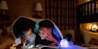 Two young boys reading a book using a torch while lying under the covers on a bed