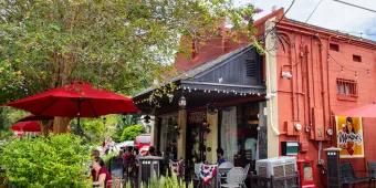 Outside of a restaurant where there are bushes to the left of a terrace are, with American circular flags and a black cover over the area. The building is orange/red.