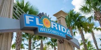 Florida welcomes you sign A blue sign with white text which says 'Florida Welcomes You' with palm trees in the background.