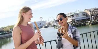 Two women stood eating snacks in Universal Studios Florida, with a lake and rollercoasters in the background. One is eating a long churro and the other is eating a pretzel.