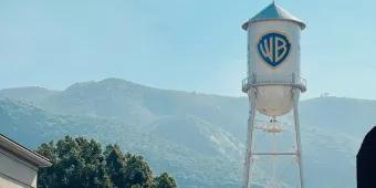 Warner Bros. Studio White water tower with blue shield shaped logo with letters WB on. In the background there is a large tree and big mountains with a blue sky overhead.