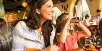 Sanaa, Disney's Animal Kingdom Villas - Kidani Village A woman and child dining in the Animal Kingdom Lodge, the mother is smiling looking off camera through a window and the little girl is also smiling looking in the same direction with binoculars held against her eyes. The mother has a bowl of seafood in front of her whereas the girl has a plate with chicken curry, rice and green beans.