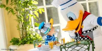 Walt Disney World Dining Reservations Donald Duck dressed as a chef leaning down to greet a child sat at a table wearing a Donald Duck hat