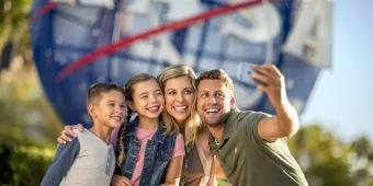 Kennedy Space Center Family of four, a man holding up a phone to take a selfie with wife and two kids, one boy and one girl. They are stood in front of a large sign, shaped like the earth, circular with red stripes over and the letters in white spelling NASA.