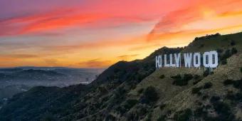 Large mountains surrounding a city. There is a large sign with capital letters spelling out "HOLLYWOOD". In the background there is a sunset with pink and orange colouring.