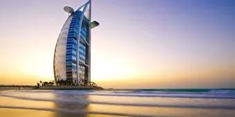 Burj Al Arab Sail shaped building coloured white with many windows and a helicopter pad on top. It is in the middle of the ocean on a man made island, with one road connecting it with mainland.