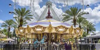 ICON Park A gold and white carousel with different pastel coloured horses and gold bars, in front of 6 palm trees diagonally lined behind and a huge ferris wheel which is white.