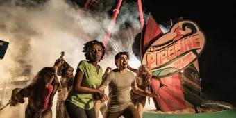 Pipeline scare zone Group of young adults running through the park with a red rollercoaster in the background and fog. Through the fog you can see a scare actor with scary zombie makeup.