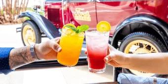 Two people clinking glasses of cocktails in front of an old fashioned car with Ford's Garage on the side
