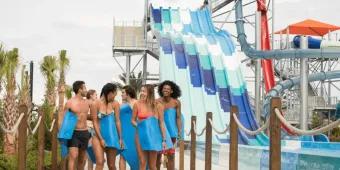 Island H2O Water Park Group of six young adults holding blue mats walking away from large blue water slides. There are six identical slides with a small drop which leads to a bigger drop.