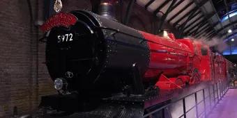 Hogwarts Express Large steam train with the label Hogwarts Express, coloured black and a dark red. There is steam surrounding and it is parked in a large factory type building.