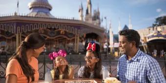 Dining Plan Family of four dining together in Magic Kingdom Park with carousel behind them. The two little girls are wearing minnie ears and smiling.