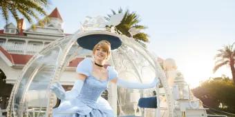 Cinderella, a blonde princess in a blue ball gown is stepping out of a glass carriage in front of a clean large white hotel.