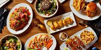 Sports & Social View from above a wooden table stacked with delicious dishes. From burger and chips, loaded nachos, pizza with cruditees and more!