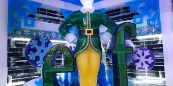 Sign of green letters saying elf in font from movie but instead of an L there is an ice sculpture of Buddy the Elf stood up straight wearing a dark green elf suit.