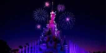 Castle lit neon pink in the night sky with purple fountains below. There are also pink fireworks in the sky.