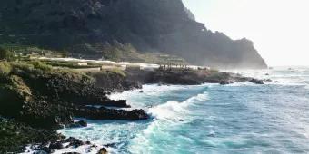 Warum Teneriffa das ganze Jahr über ein perfektes Reiseziel ist Türkisblaues Wasser wirft Wellen auf eine Küste mit Bergen im Hintergrund