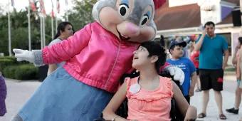 Give Kids The World A young girl in an electric wheelchair looking up at a large bunny dressed in a pink jacket and pink bow.